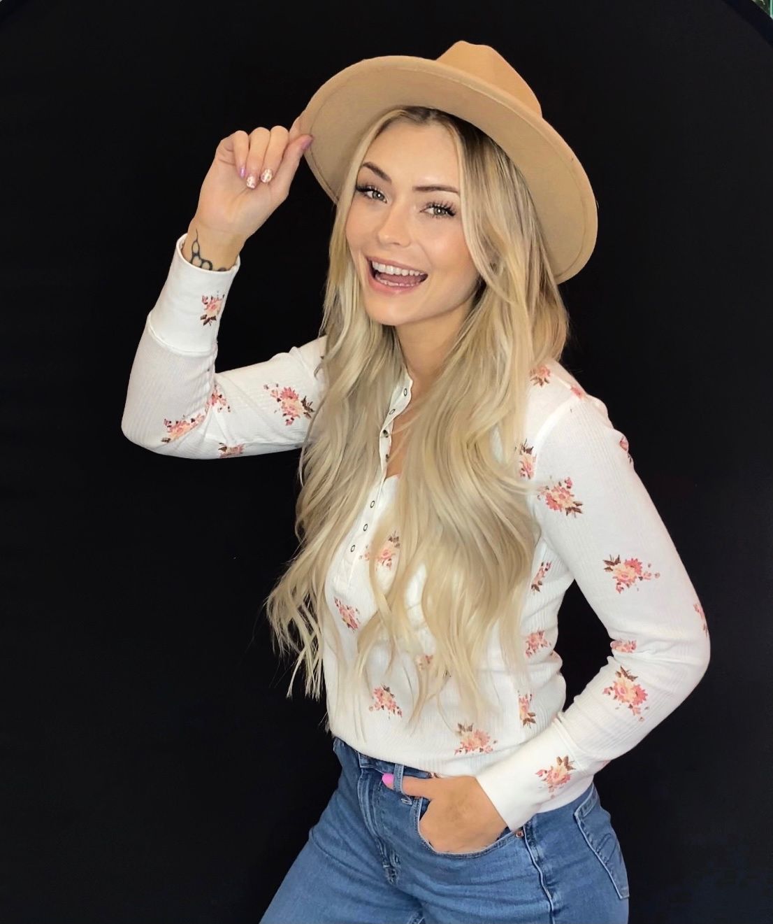 Blonde woman in hat smiles, hand on hat, wearing floral shirt and jeans, dark background.