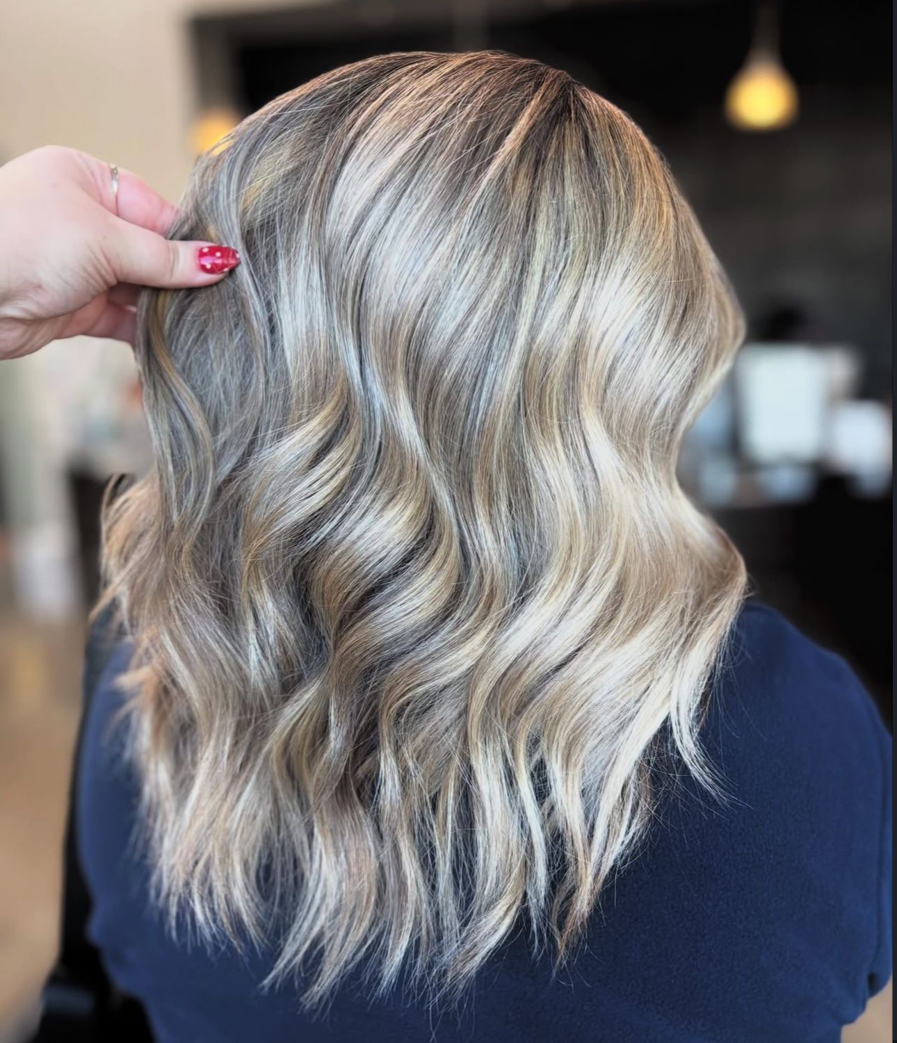 Woman with wavy, highlighted blonde hair, styled in a salon. Hand is touching the hair.
