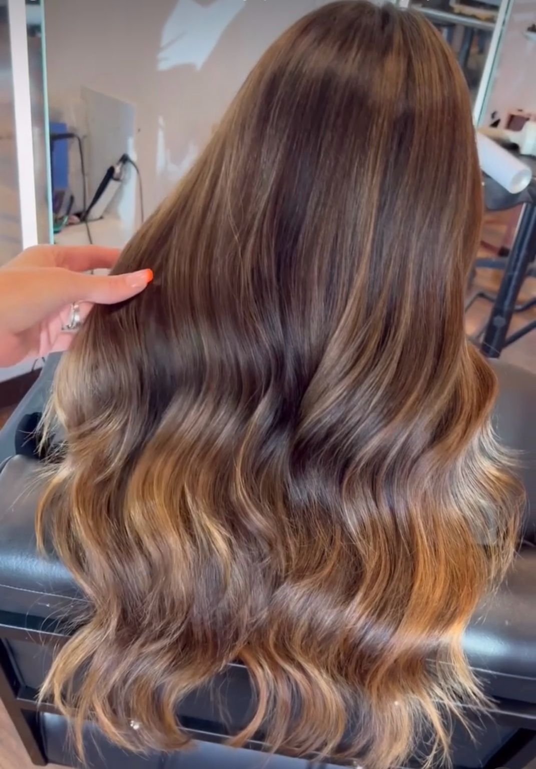 Woman with long, wavy brown hair, highlighted with lighter tones; hand touches the hair in a salon.