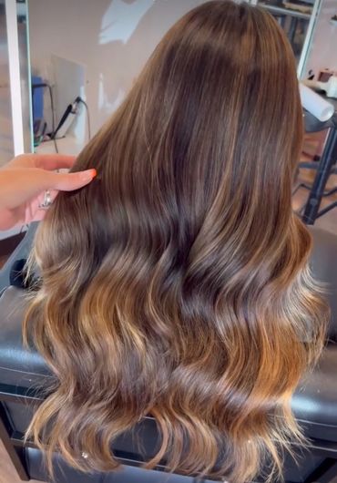 Woman with long, wavy brown hair, highlighted with lighter tones; hand touches the hair in a salon.