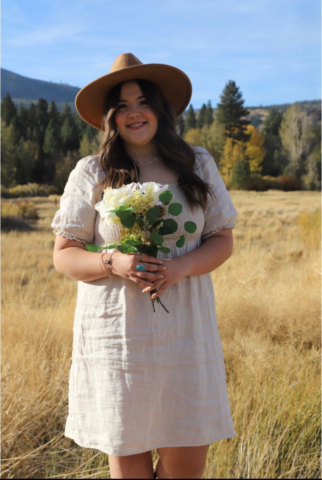 Woman in dress and hat holds flowers in a field with mountains and trees in the background.