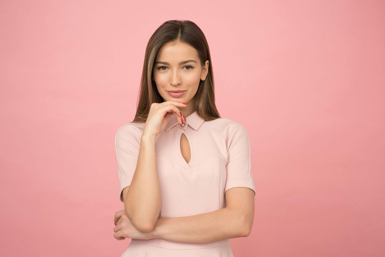 Woman in pink dress with hand on chin, smiling at the viewer against a pink backdrop.