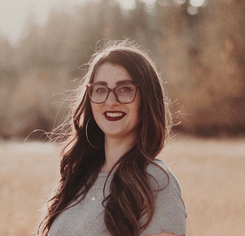 Woman with long brown hair and glasses smiles outdoors, wearing a grey shirt, field background.