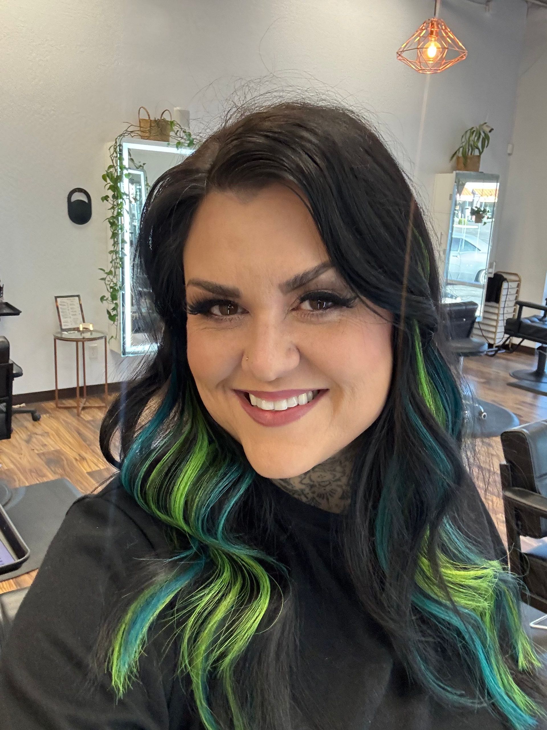 Woman with black hair, teal and green streaks, smiling at the camera in a salon.