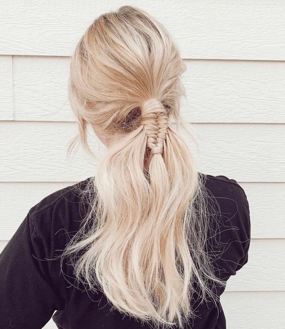 Blonde hair in a low ponytail, with a small braid holding it together. Against a white wall.