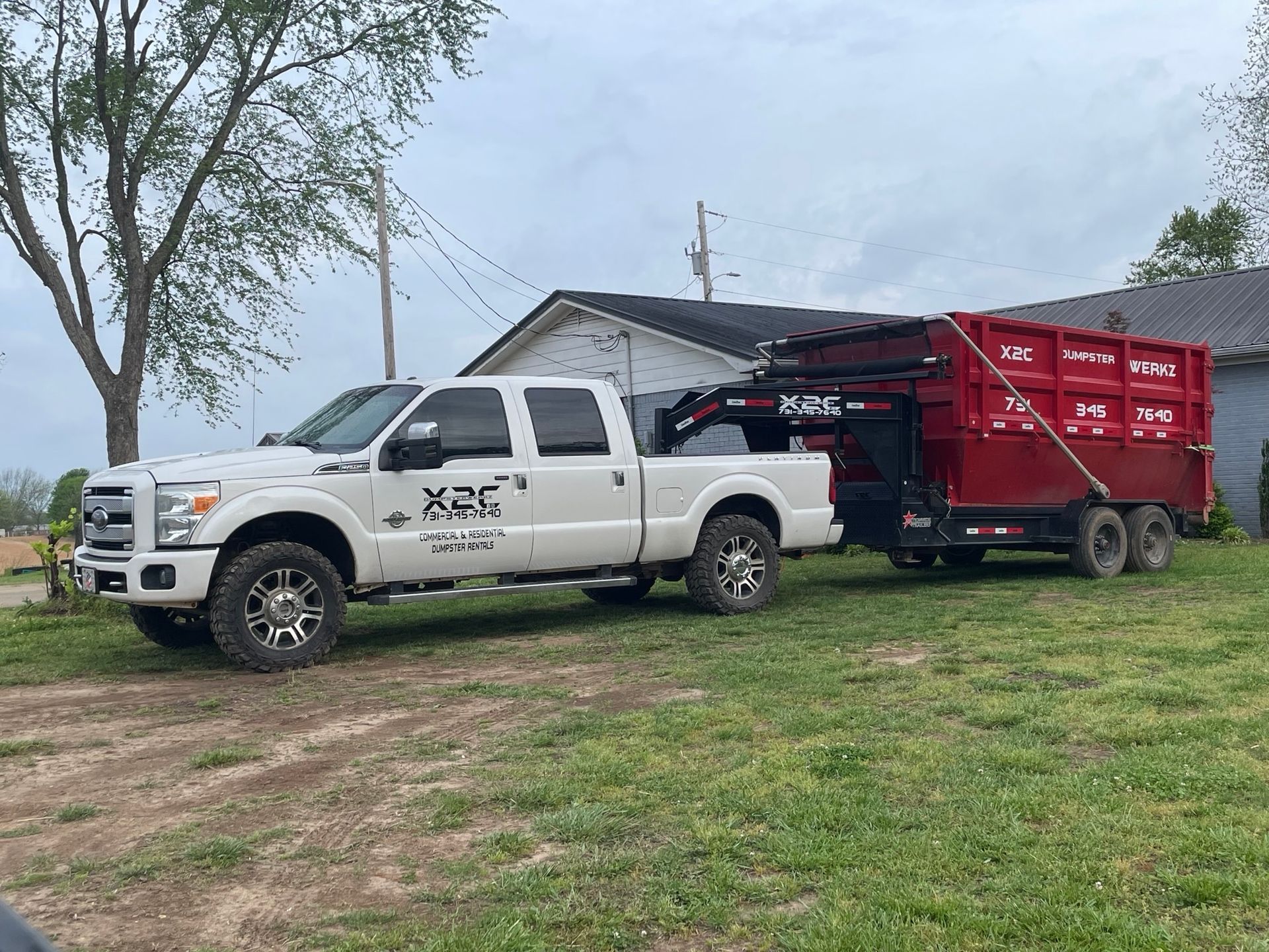 A white truck is towing a red dumpster in a grassy field.