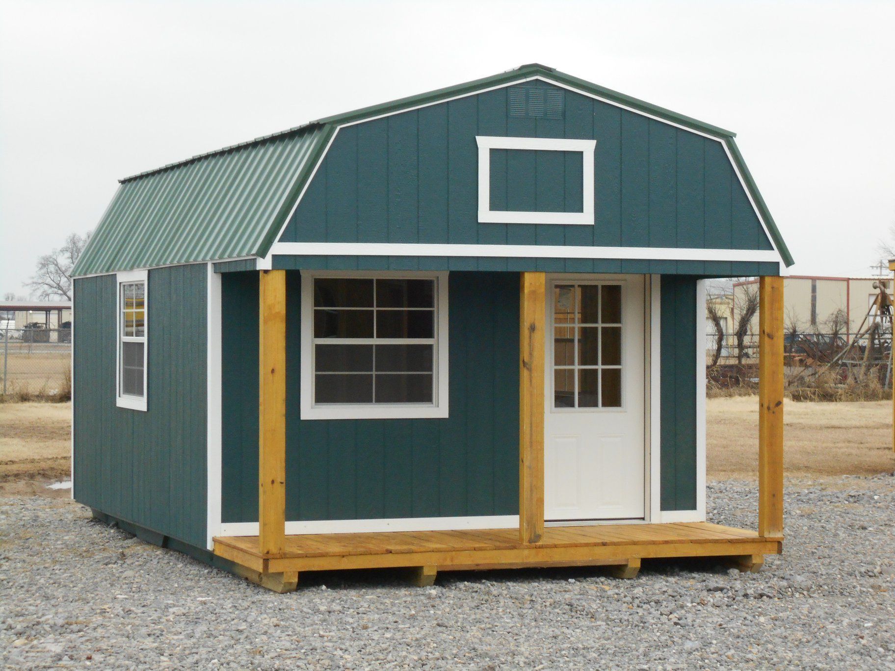 Green shed with porch and white trim. Gravel ground and a metal roof.