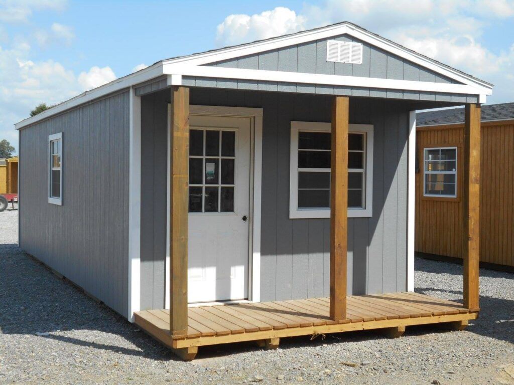 Gray shed with a wooden porch, white door and windows, brown support beams, and gravel ground.