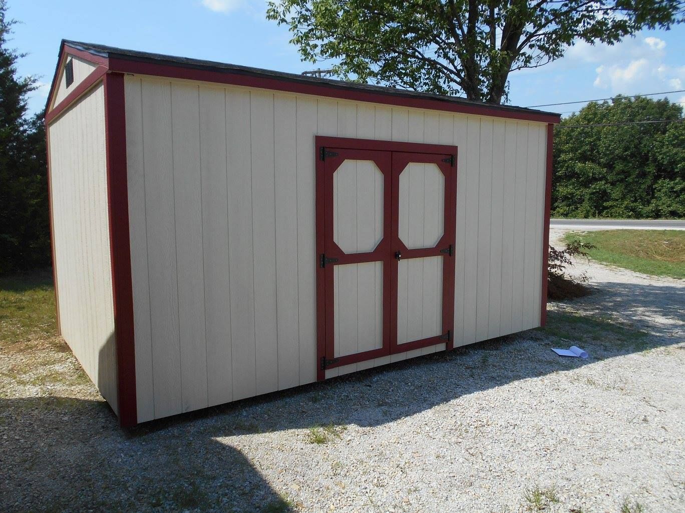 Tan shed with maroon trim and double doors on gravel.