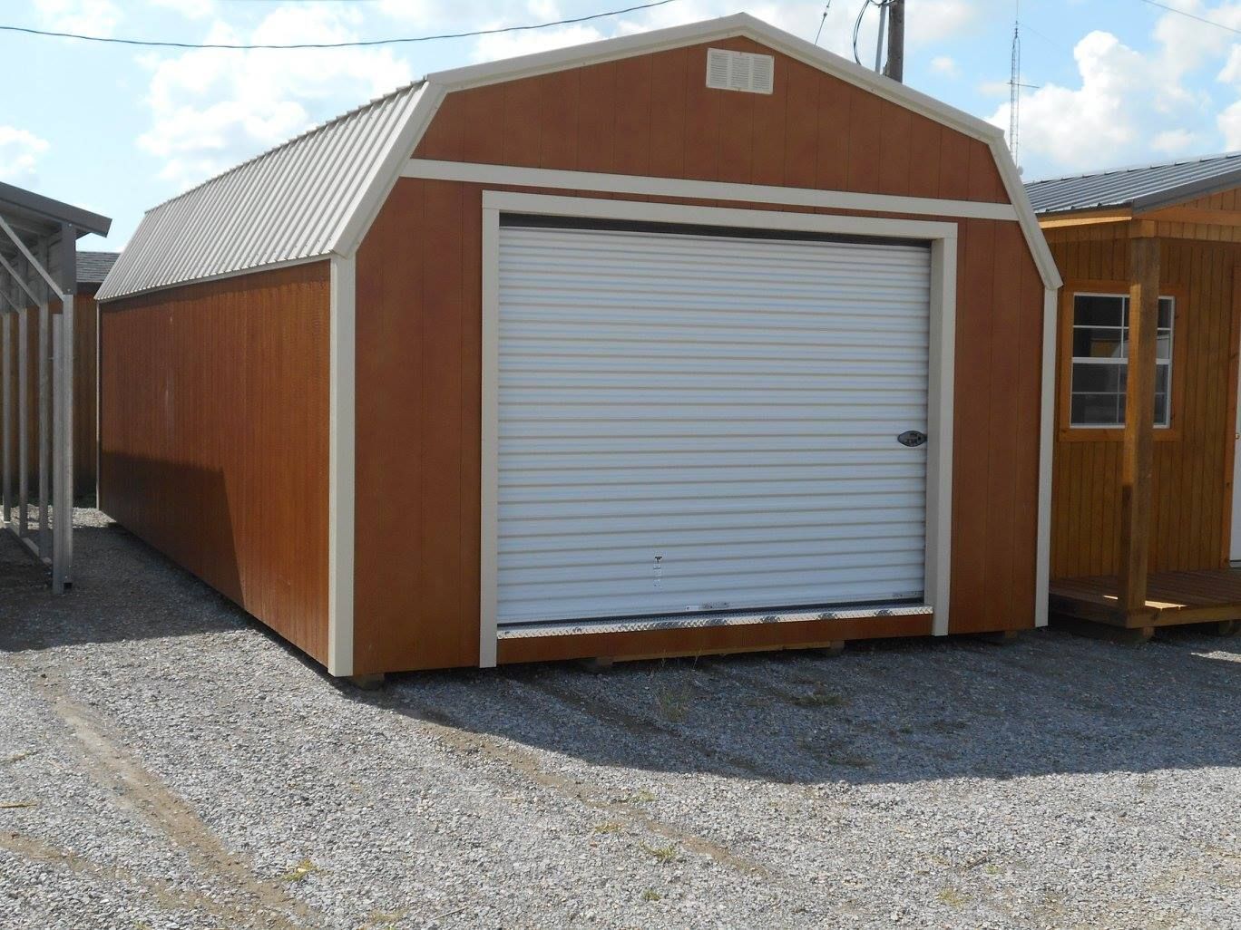 Brown and white barn-style shed with a large garage door on a gravel lot.