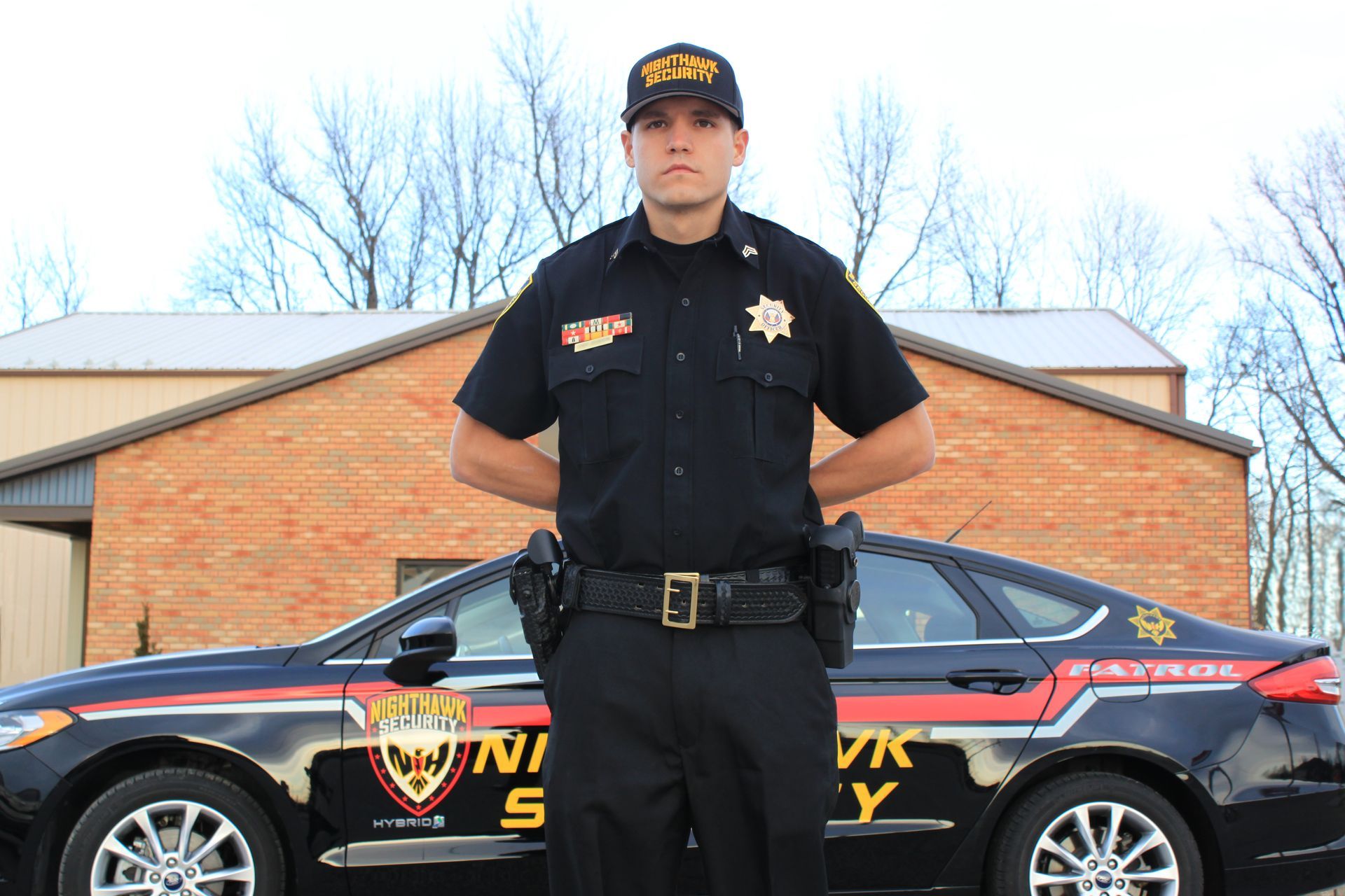 Law enforcement officer in dark uniform stands by patrol car. Building and trees in the background.
