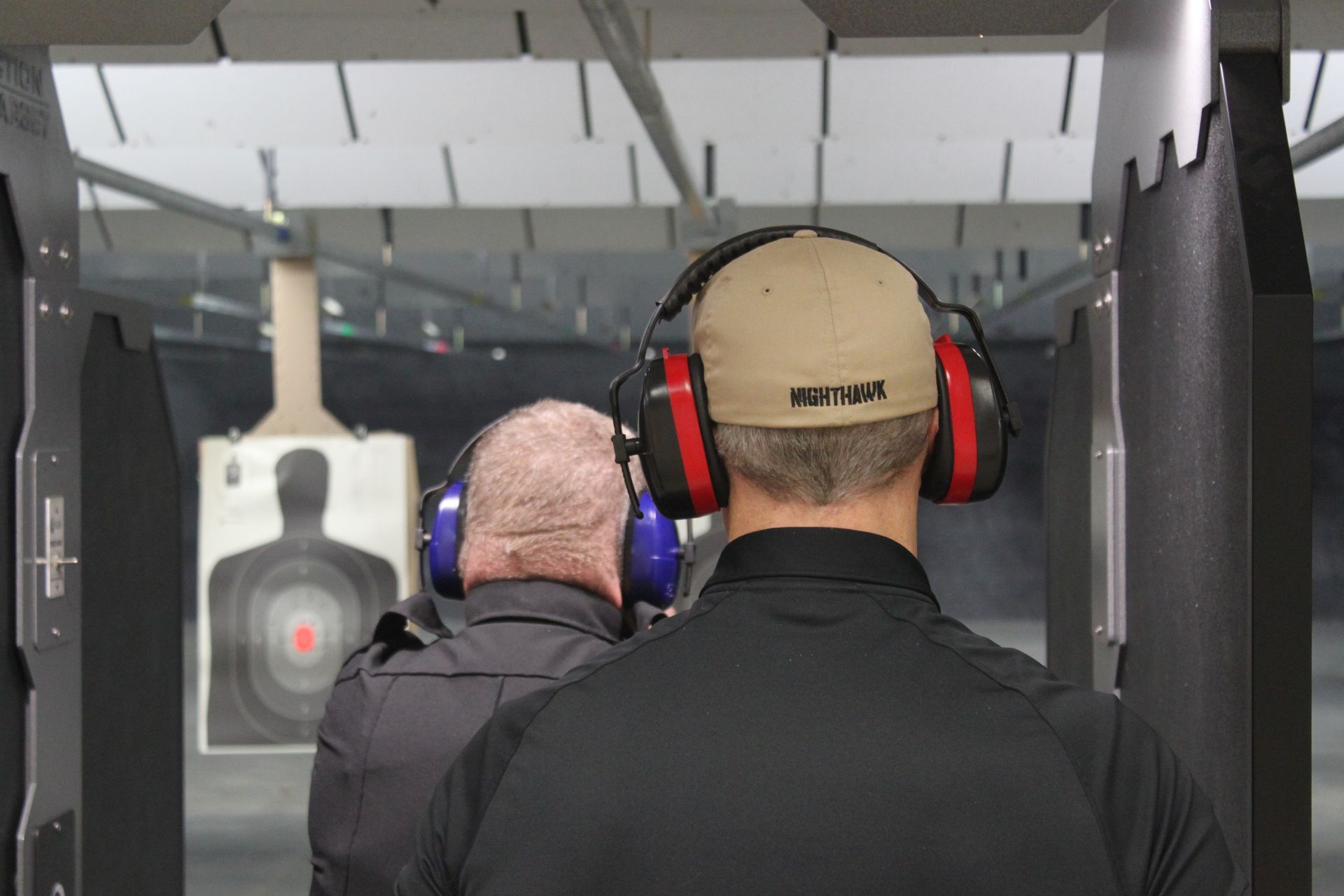 Two people at an indoor shooting range, wearing ear protection, aiming at targets.