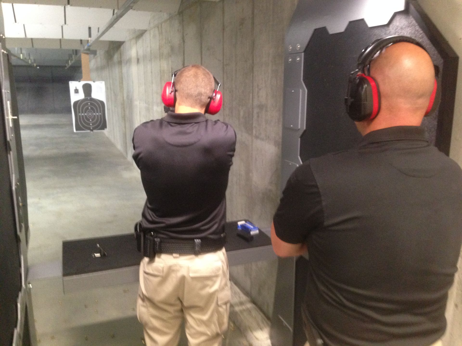 Two men at an indoor shooting range; one aiming at a target, the other observing. Both wear ear protection.