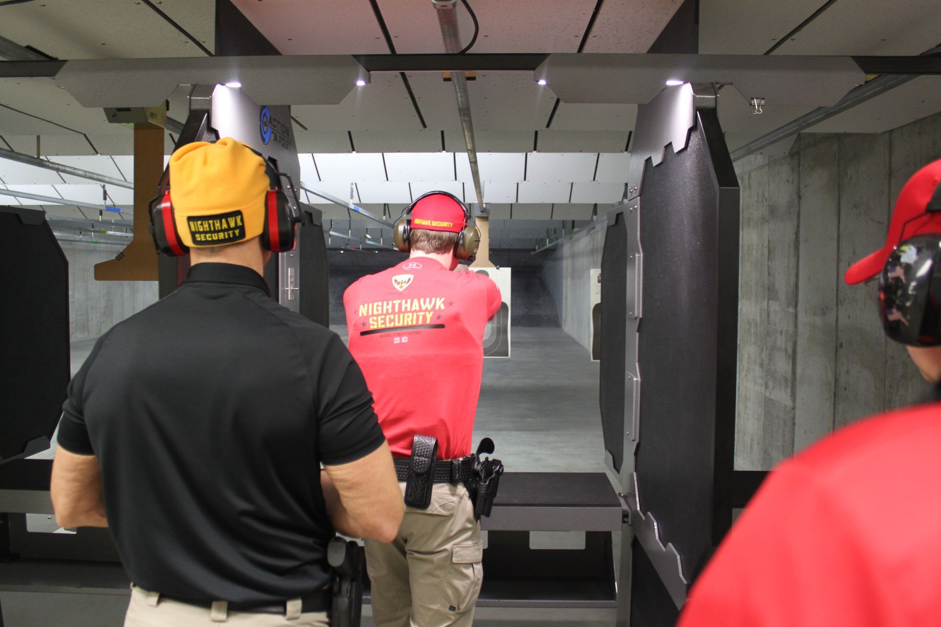 People at a shooting range; one aims a pistol. Others observe, wearing ear protection and hats.