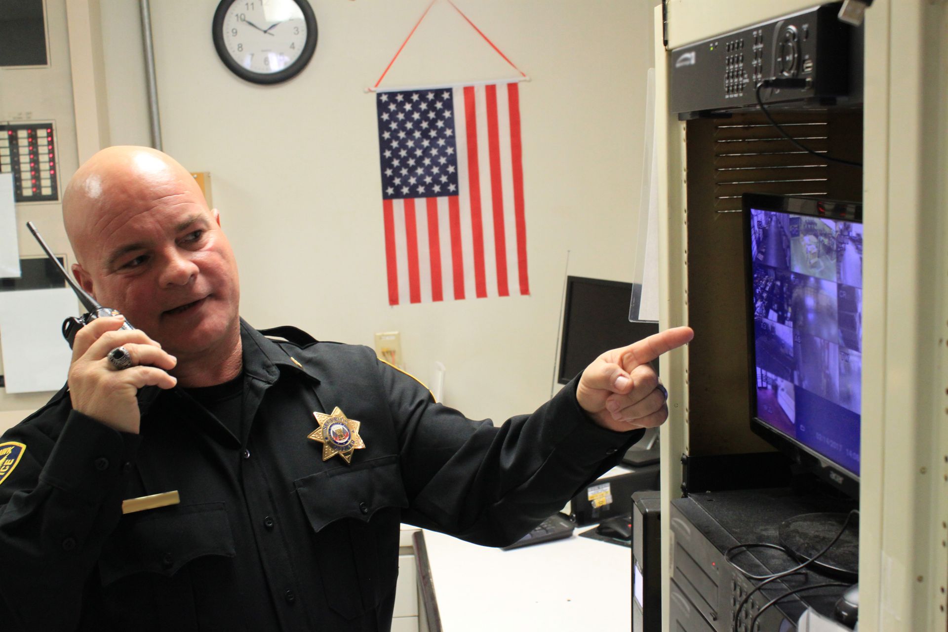 A uniformed security officer points at a monitor with surveillance footage, holding a walkie-talkie. US flag visible.