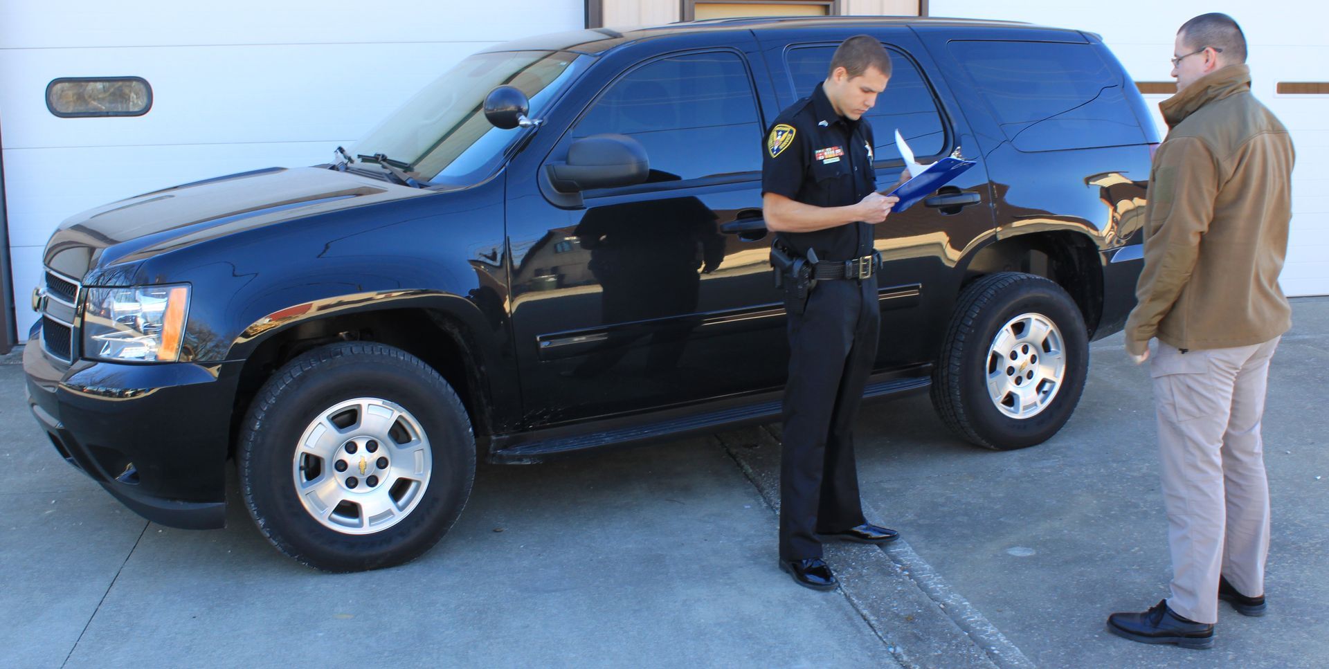 A police officer writes on a clipboard next to a black SUV, talking to a man.
