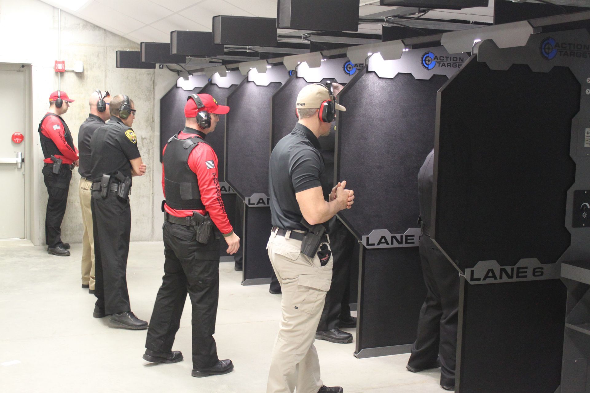 People in a shooting range, some aiming, others observing. Red hats and vests, black booths and targets.