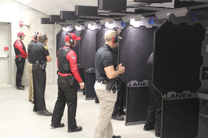People in a shooting range, some aiming, others observing. Red hats and vests, black booths and targets.