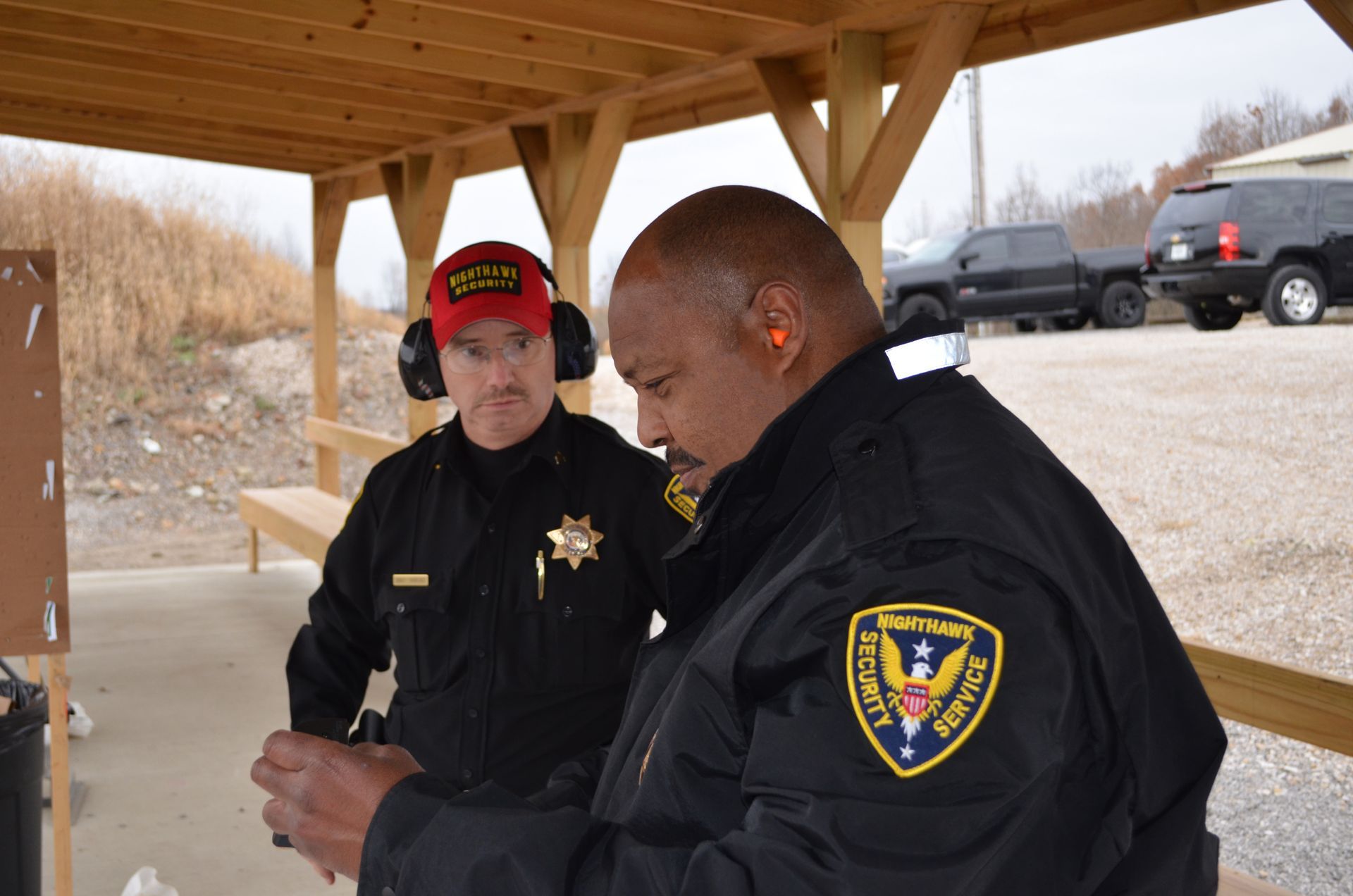 Two men at a shooting range, one examining a handgun. One wears a badge. They are outdoors under a wooden shelter.