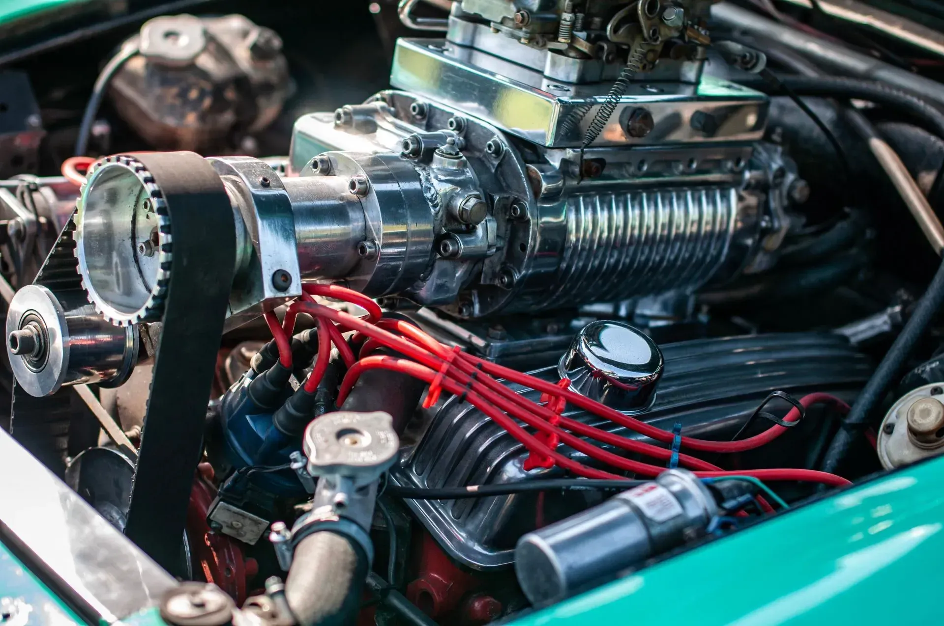 A close-up of a bright teal car's powerful engine with a prominent chrome supercharger and red ignition wires.