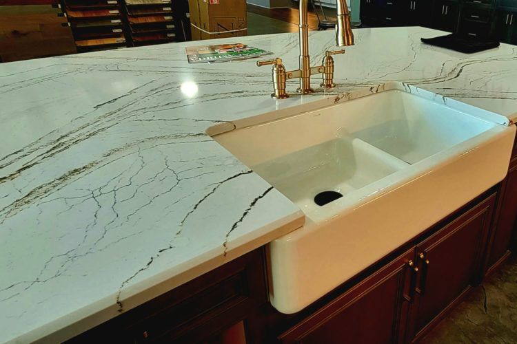 White farmhouse sink in a marble-patterned kitchen countertop with brass faucet and dark wood cabinets
