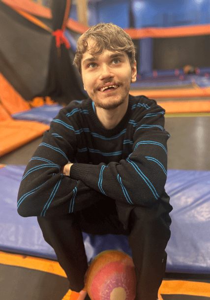 A man is sitting on a trampoline with his arms crossed and holding a ball.