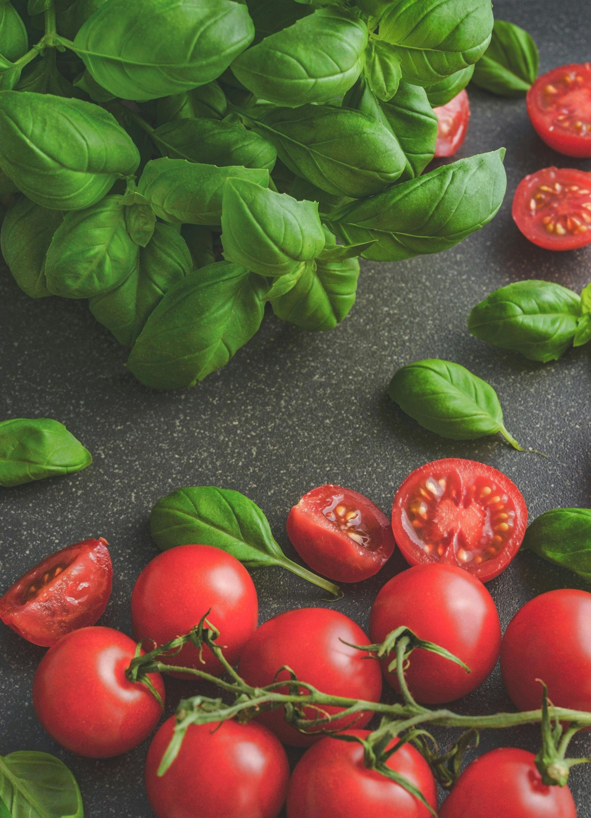 Photo of a grey surface with bright red tomatoes and green basil leaves