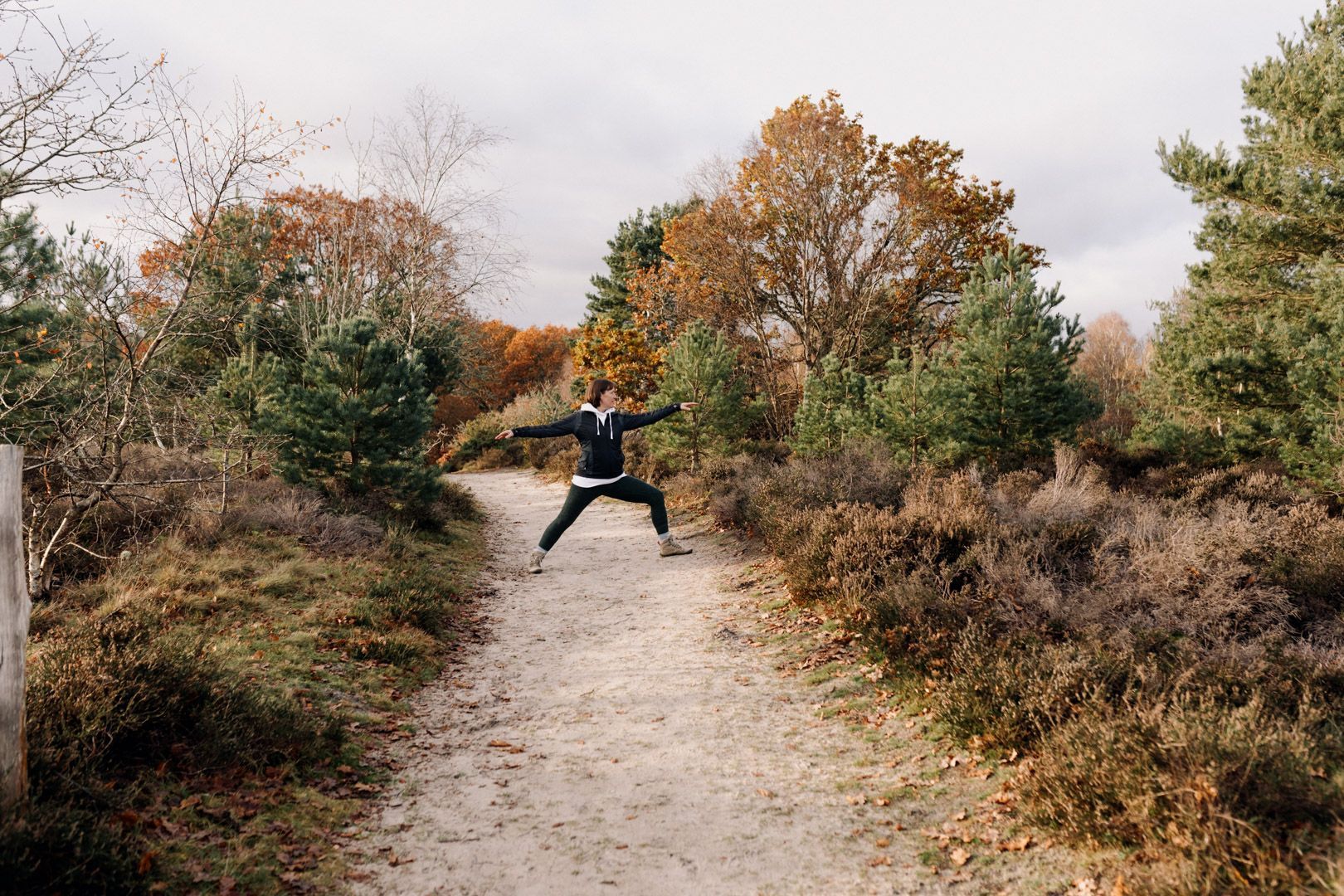 photo of a lady doing a yoga warrier pose in the autumn on a sandy path lined with bushes