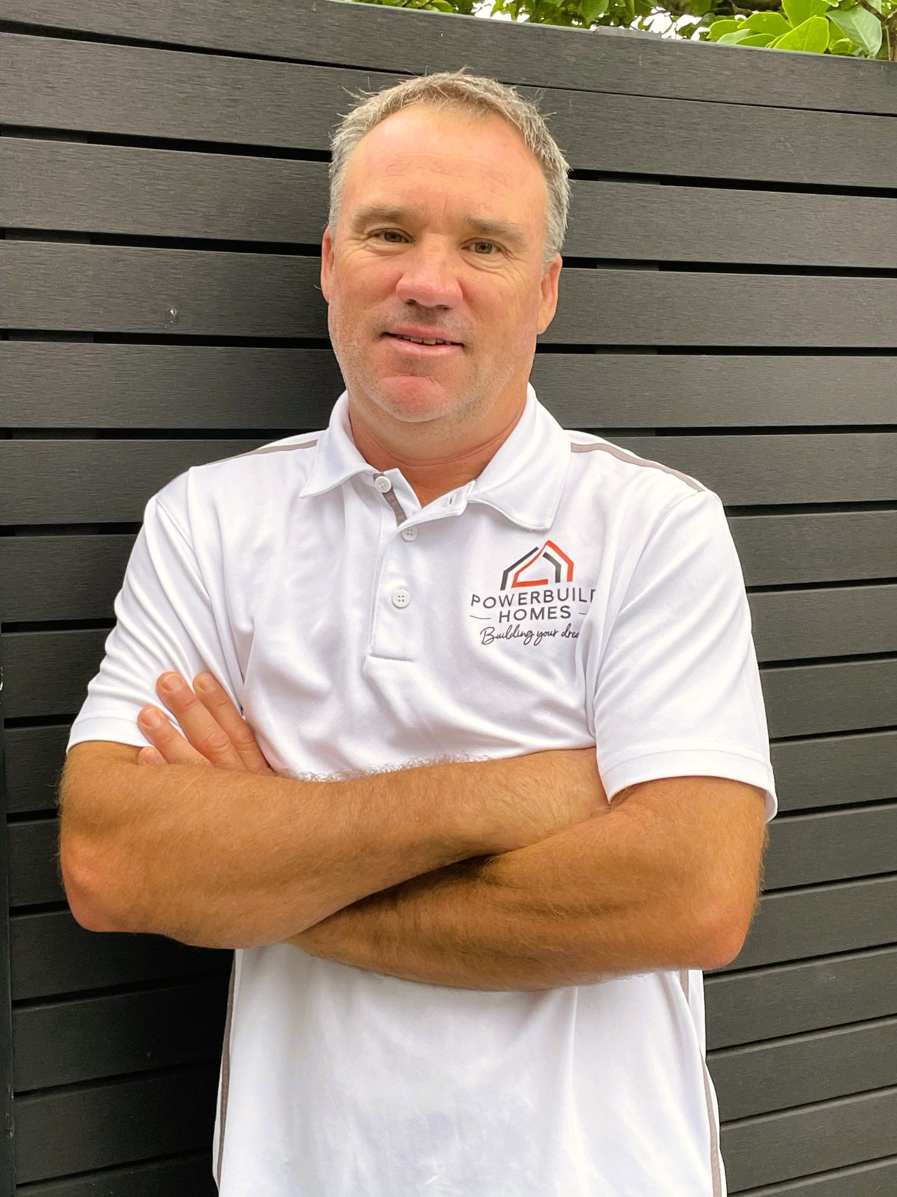 Man in white polo shirt with crossed arms stands in front of a dark gray wall.