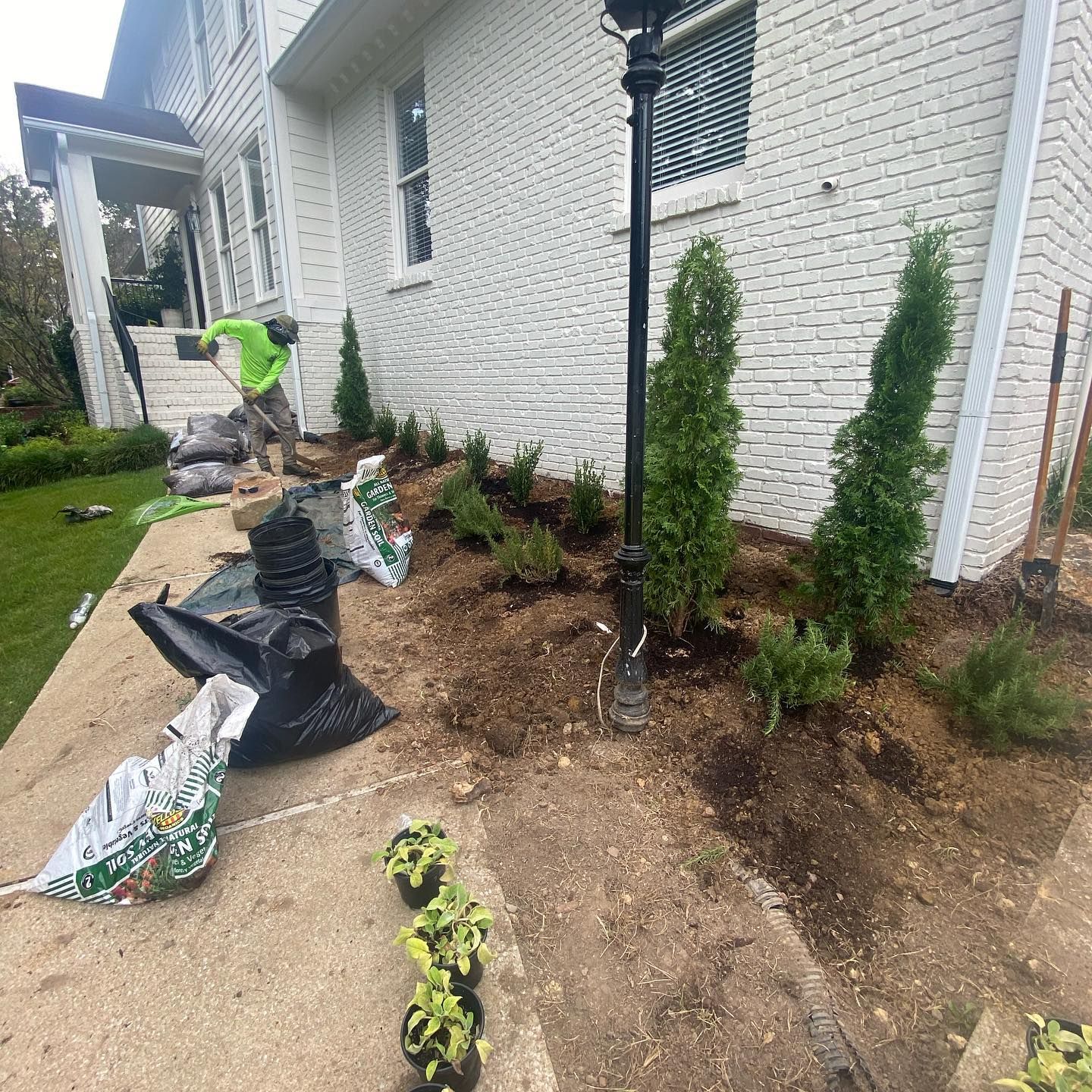 A man is planting trees in front of a white brick house.