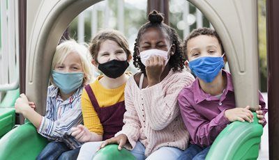 Four children wearing face masks sit on playground equipment, smiling.