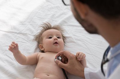 Doctor listening to infant's heart with stethoscope on a white bed. Baby looks up, arms raised.