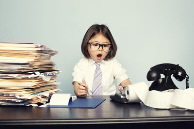 Child wearing glasses and tie, with a surprised expression, sits at a desk piled with papers and a calculator.