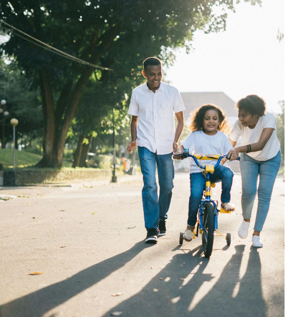 Family walking and biking in a park; child rides a bike with training wheels, parents nearby.