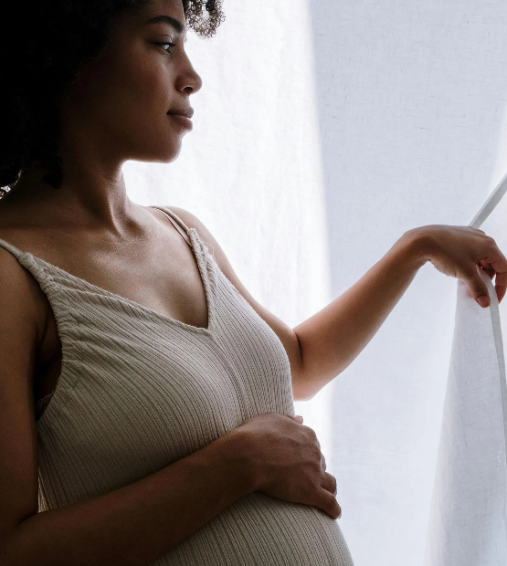Pregnant person in a cream-colored dress, gazing out a window, hand on her stomach.
