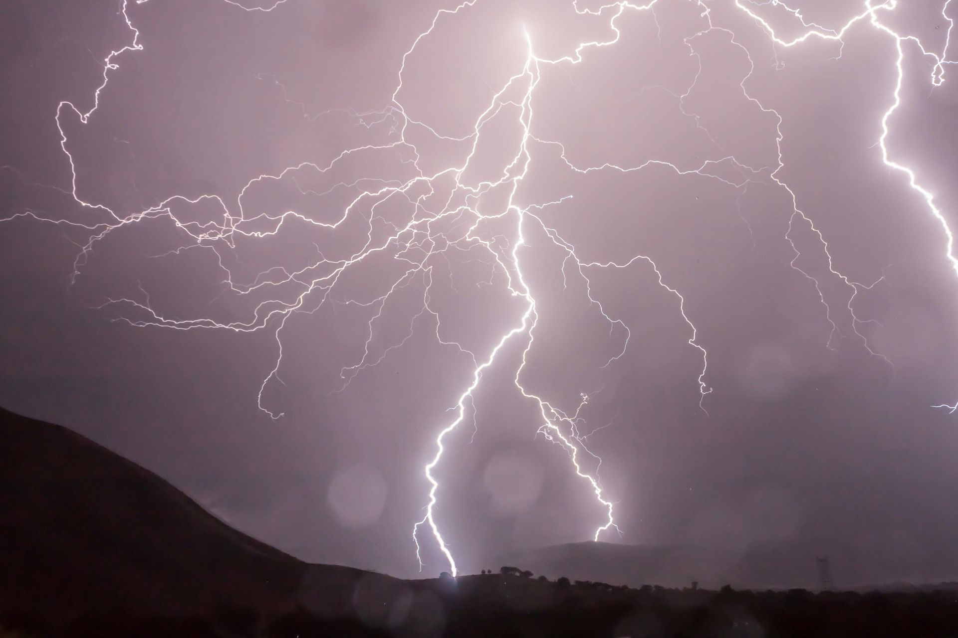 Lightning strikes over a dark landscape, illuminating the sky with bright white flashes.