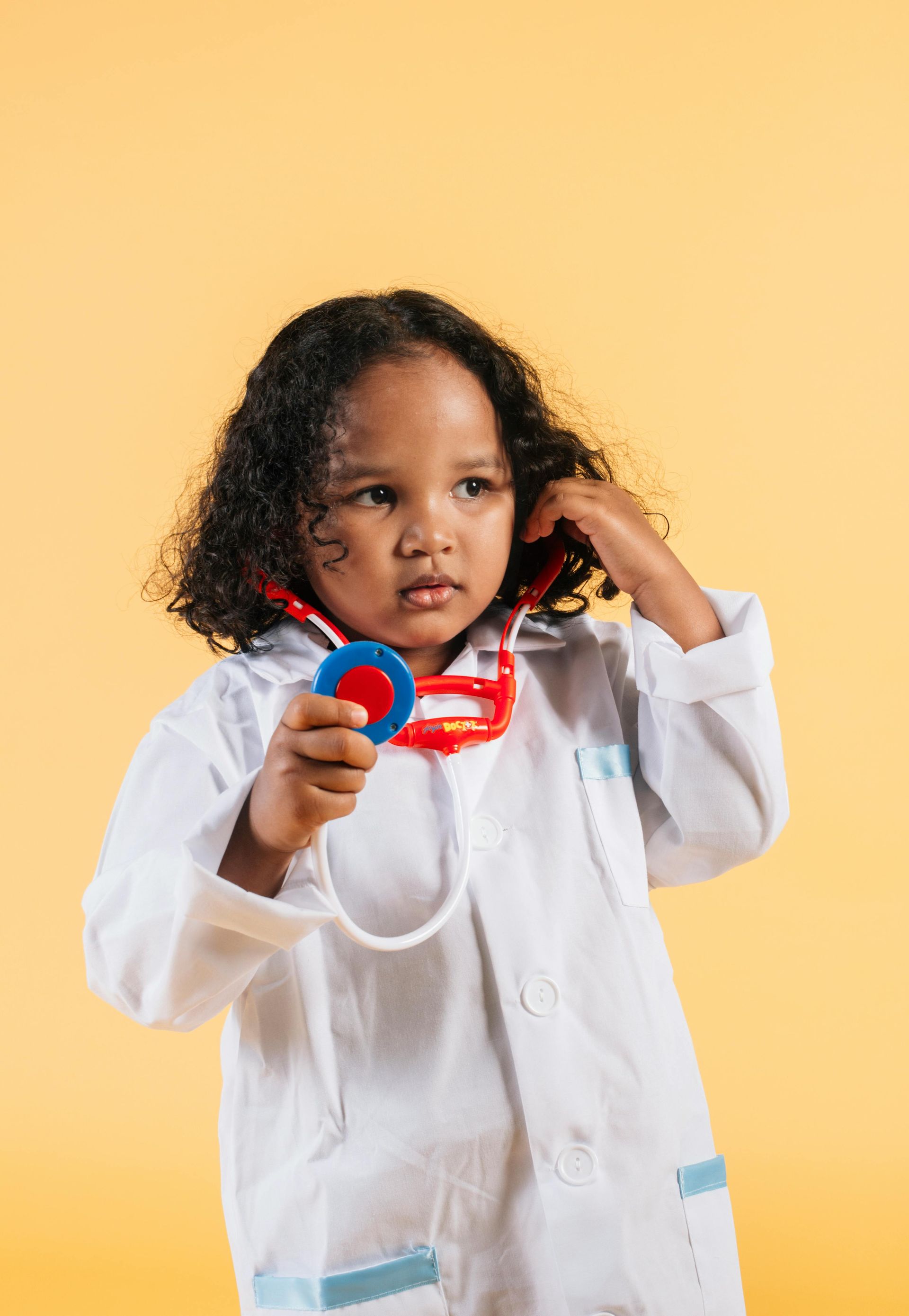 Child in a lab coat and stethoscope against a yellow background.