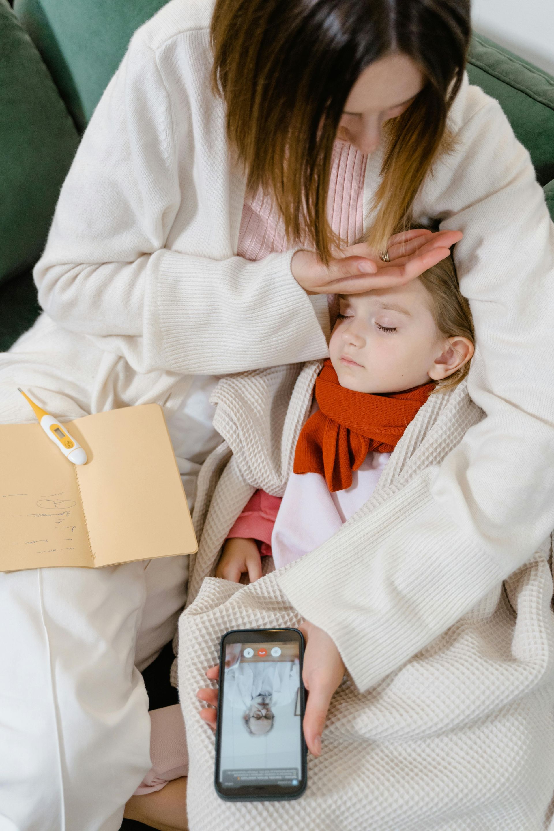 Woman checking child's forehead while holding phone showing a medical image. Child is wrapped in blanket and wearing a scarf.