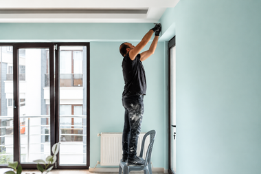 Person painting a light blue wall near a ceiling in a bright room with a ladder and large windows
