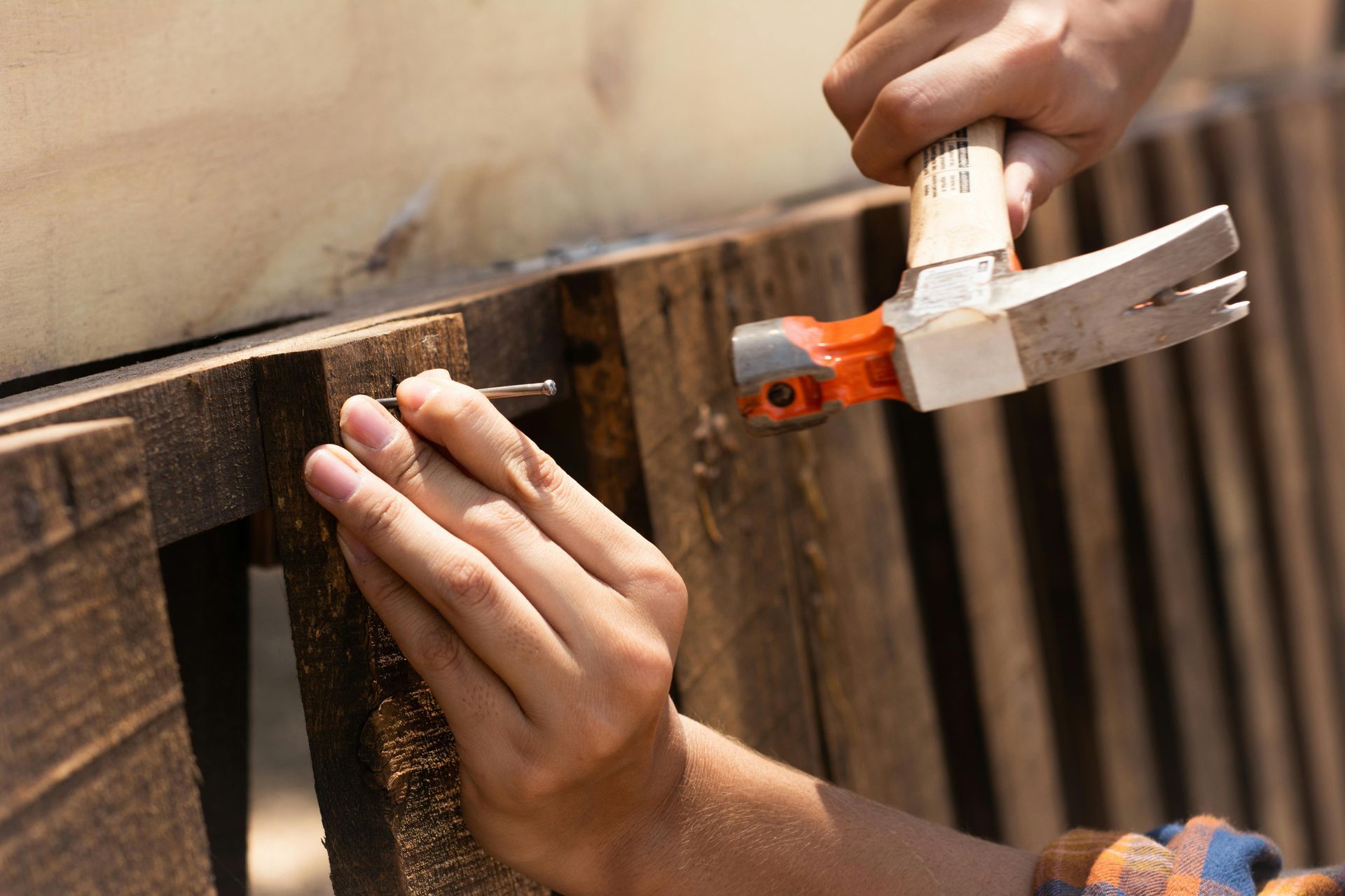 Hands using a hammer and nail on a wooden fence rail, with a small clamp holding the wood in place.