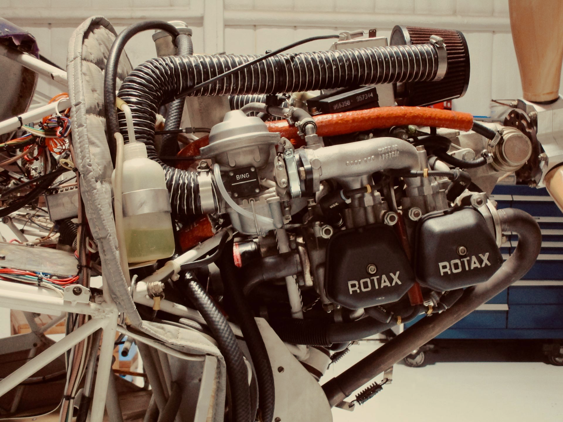 Close-up of a Kitfox aircraft with exposed Rotax engine, showing dual valve covers, Bing carburetor, air ducts, fuel lines, and tubular frame in a hangar.

