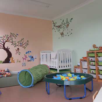 Nursery decorated in pink and mint green with tree motifs on the wall, a white cot in the corner, and lots of soft toys and toys suitable for toddlers