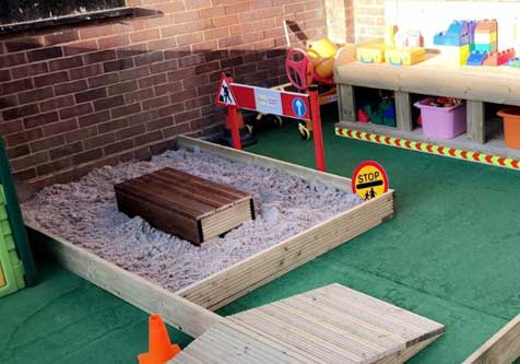 A sand pit in a play area with various blocks and other toys on a table to one side and a brick wall behind it