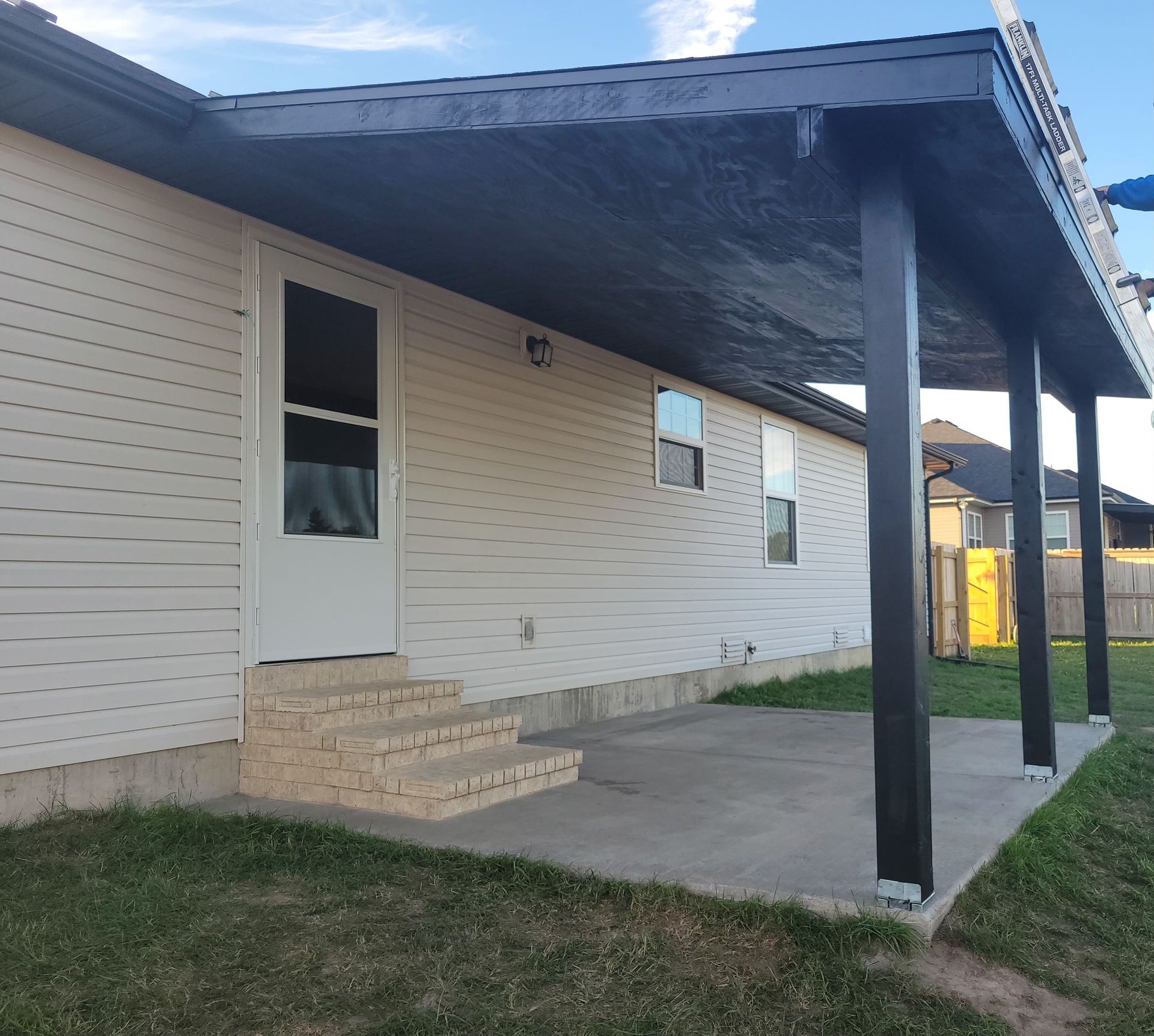 Backyard patio with a covered roof, concrete slab, brick steps, and a white door.