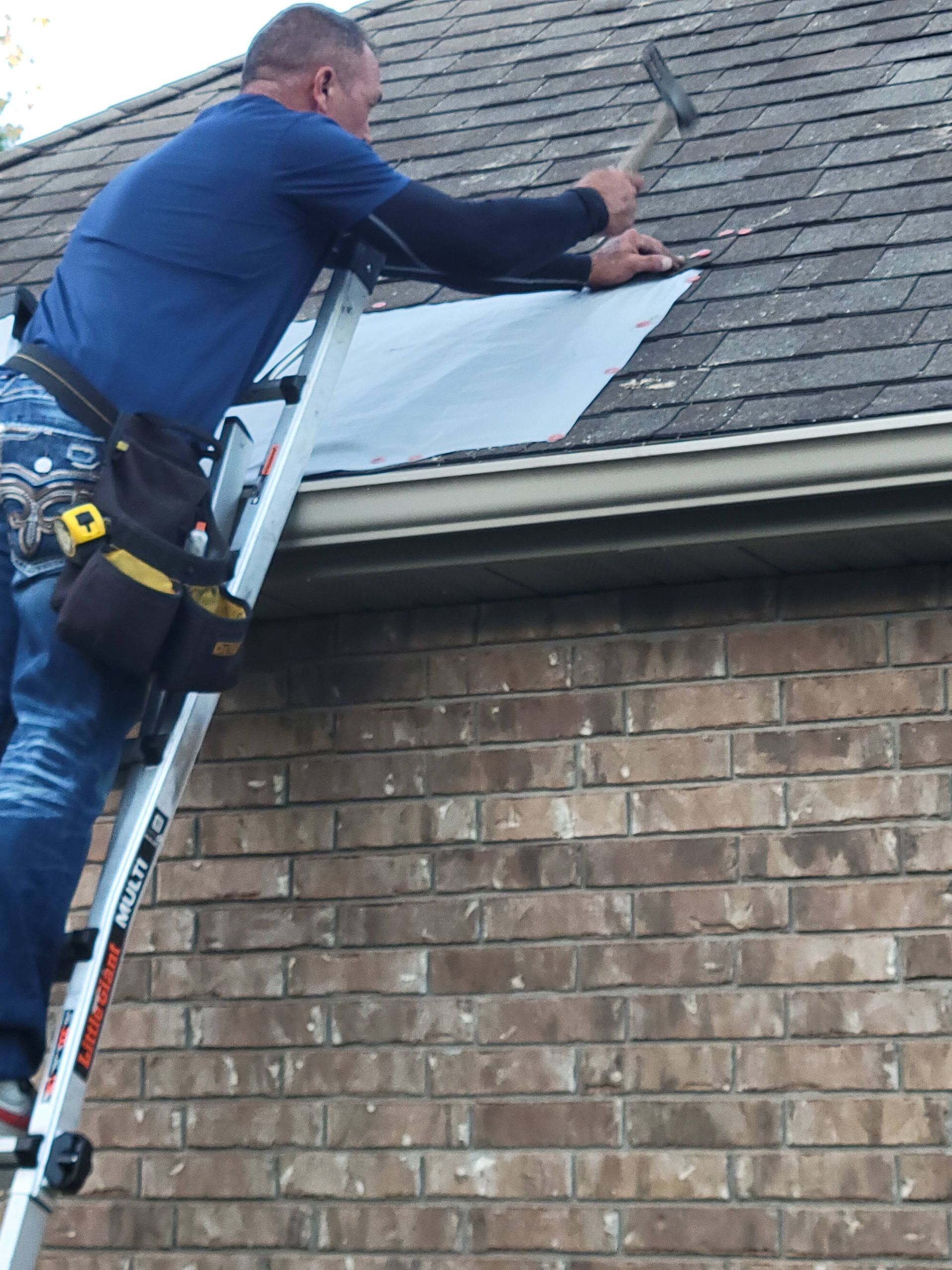 Person on a ladder hammering a dark, rectangular sheet onto a shingled roof, over a brick wall.