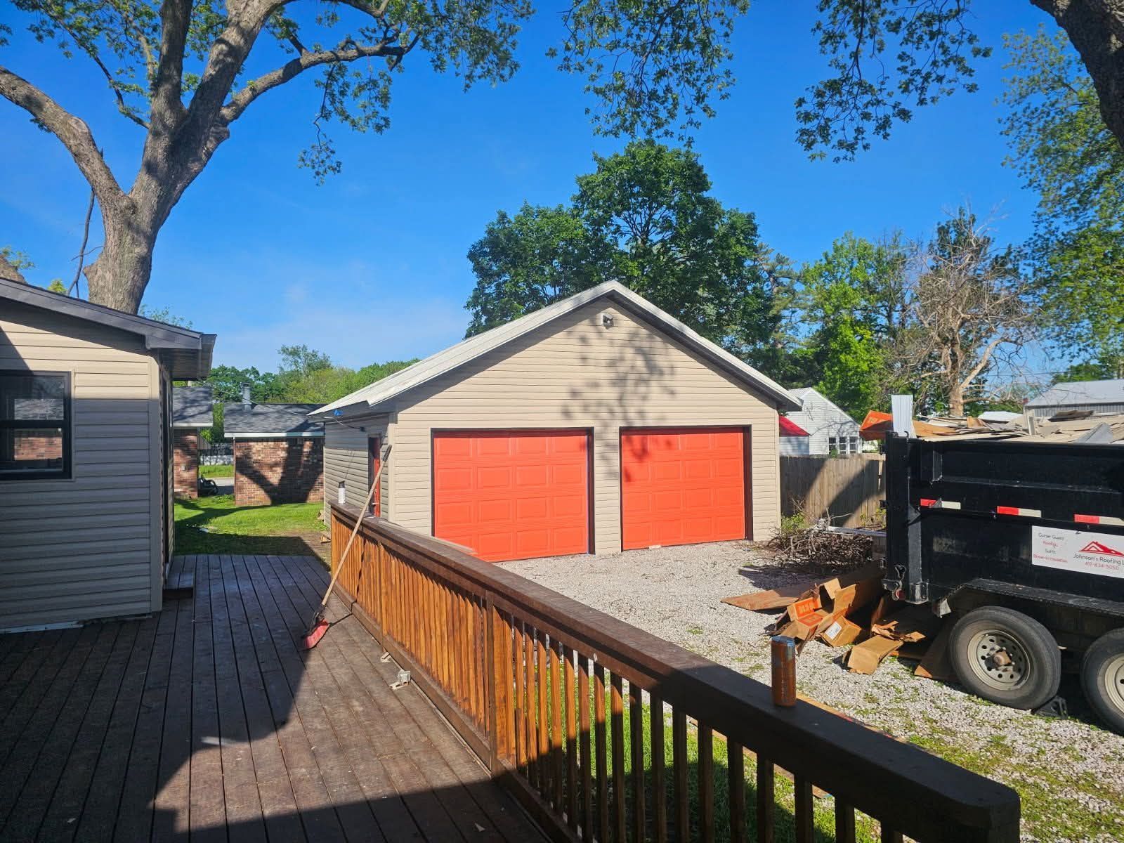 Garage with two red doors, light tan siding, gravel driveway, and a wooden deck.