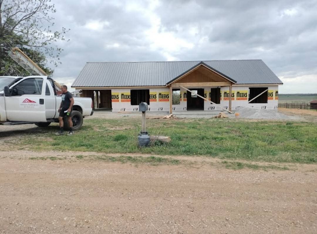 House under construction; metal roof, worker by white truck, cloudy sky, rural setting.