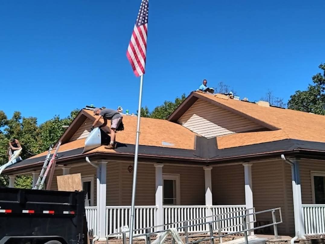 Roofers working on a house under a bright blue sky, American flag in foreground.