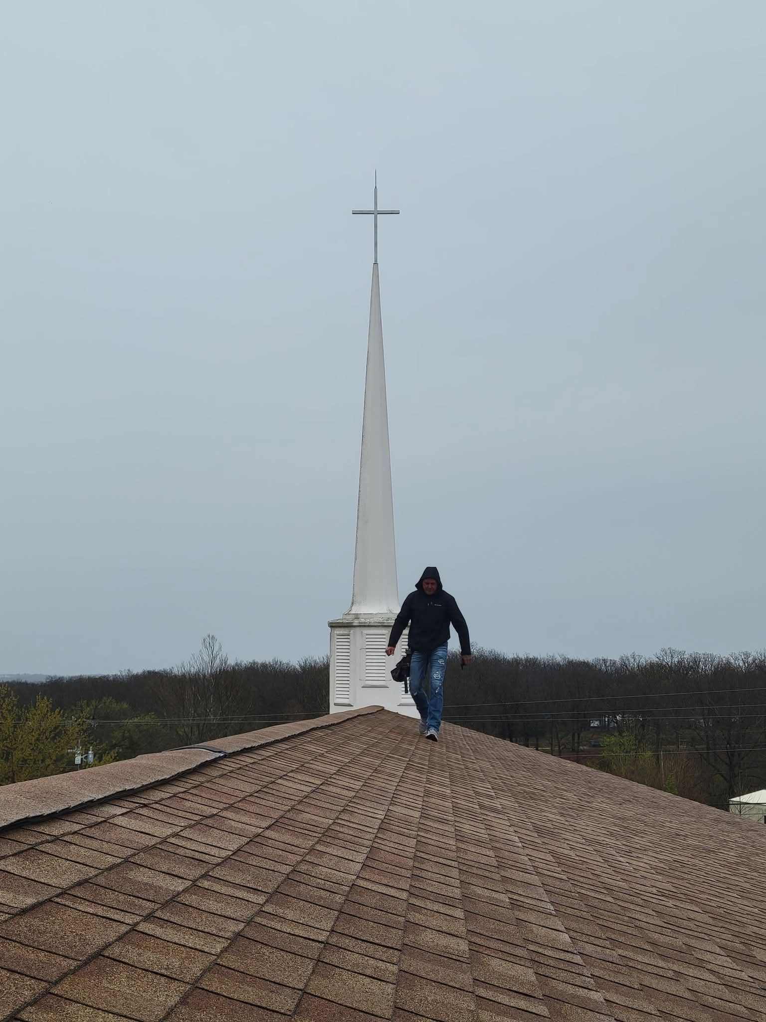 Person walks on church roof with spire and cross against cloudy sky.