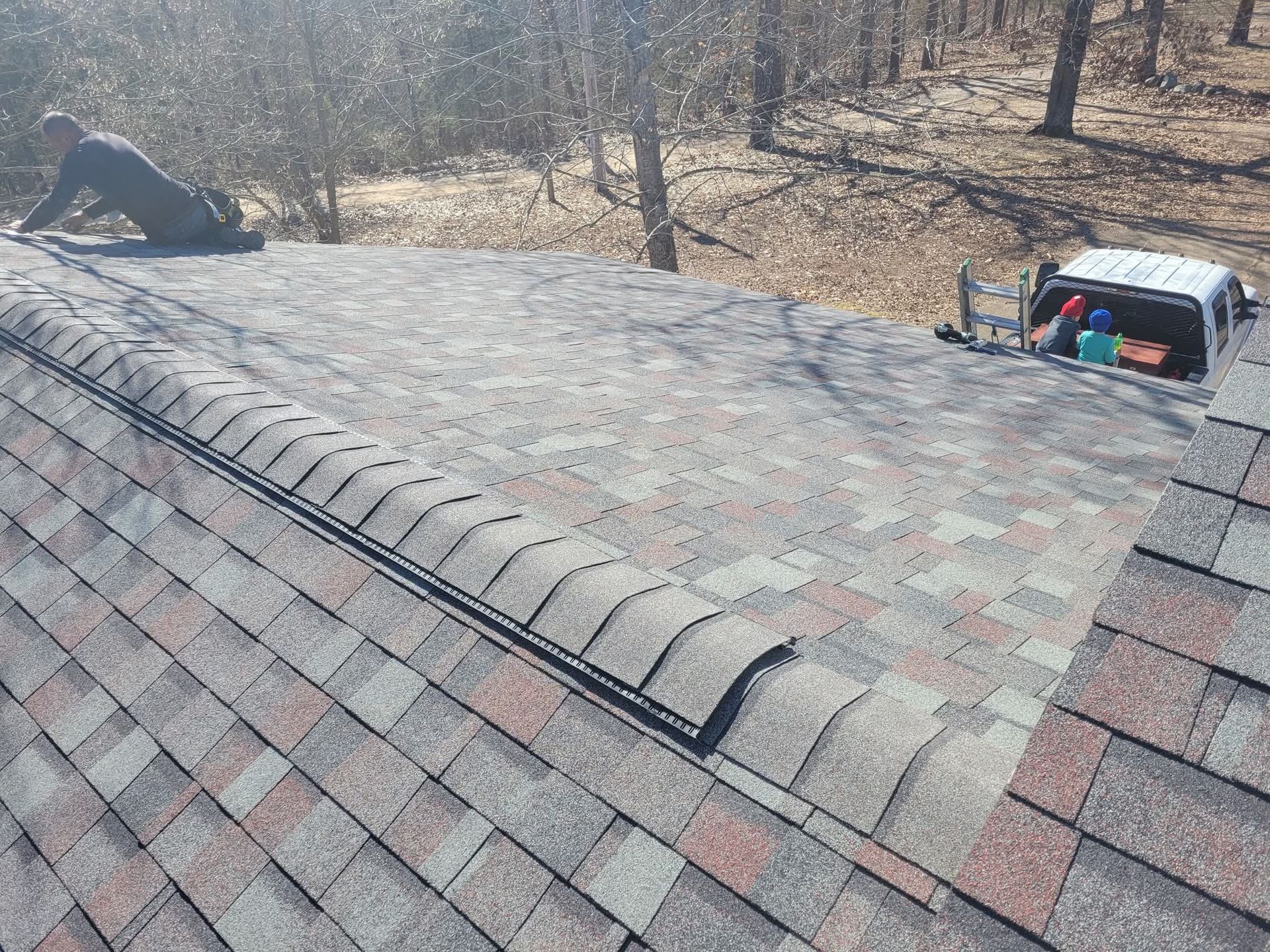 Roofer on a shingled roof near a treeline. Tools and supplies sit nearby. Sunny day.